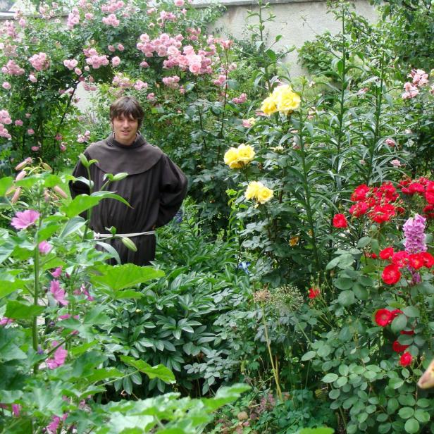 Ein Mann in einer braunen Kutte steht in einem üppigen Garten voller Blumen.