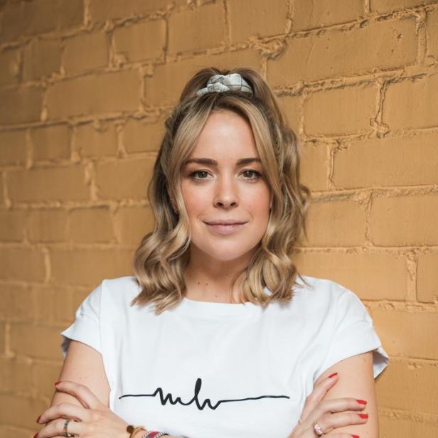 Eine Frau mit blonden Haaren und einem weißen T-Shirt posiert vor einer gelben Backsteinmauer.