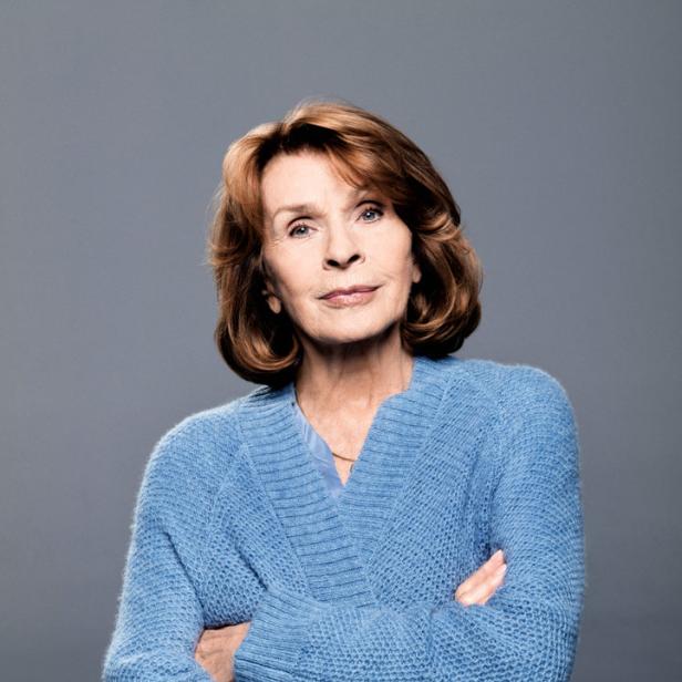 Eine ältere Frau mit verschränkten Armen trägt eine blaue Strickjacke.