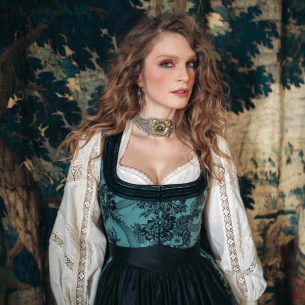 Eine Frau in Tracht vor einem Wandteppich mit Baumdarstellung.