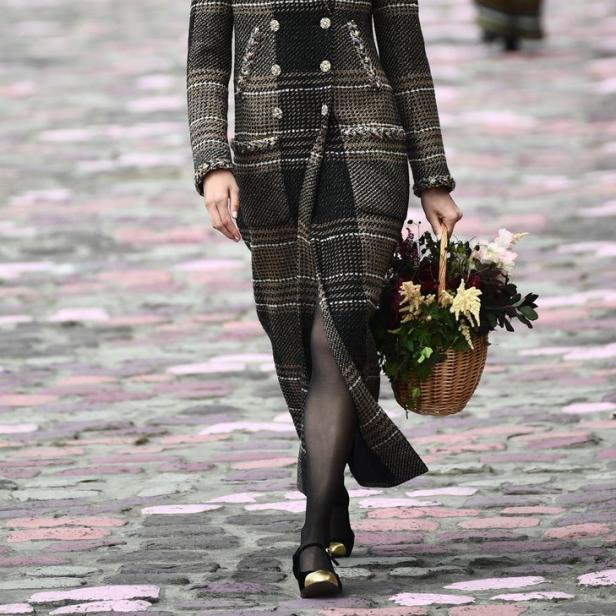 Ein Model mit einem karierten Mantel und einem Blumenkorb auf einem Laufsteg.