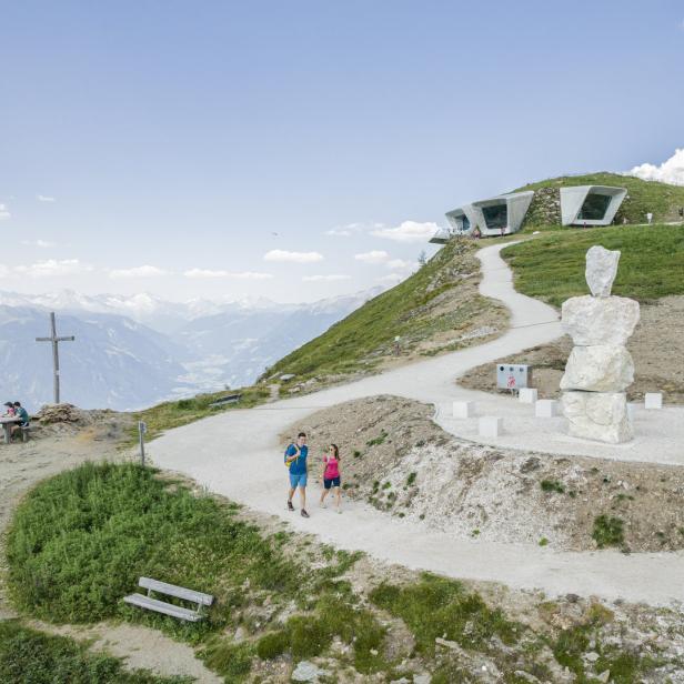 Ein Paar wandert auf einem Bergpfad mit Blick auf ein modernes Gebäude und eine Berglandschaft.