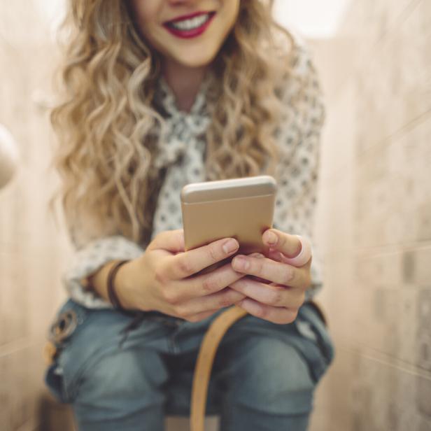 Junge blonde Frau mit langen Haaren sitzt mit dem Handy auf der Toilette.