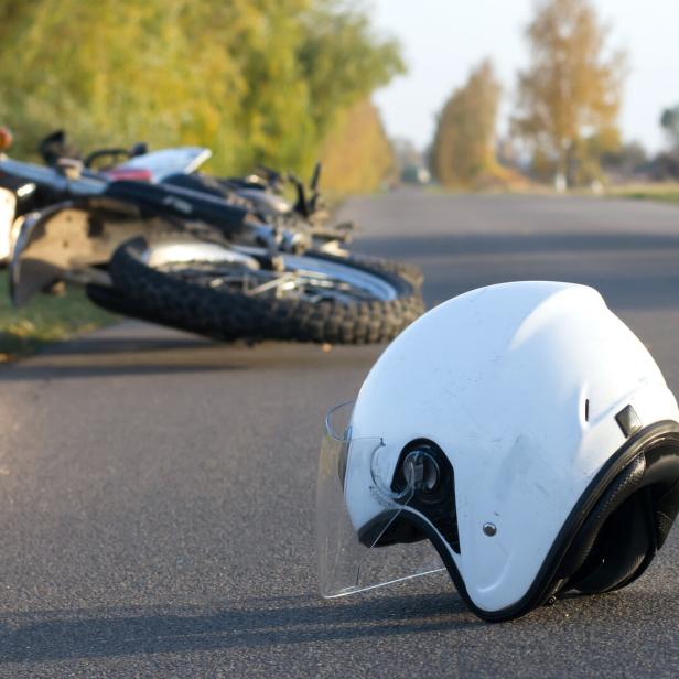 Ein weißer Motorradhelm liegt auf einer Straße, im Hintergrund liegt ein umgefallenes Motorrad.