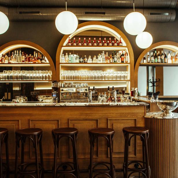 Eine Bar mit hölzernen Hockern und Regalen voller Flaschen und Gläser.