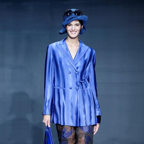 Eine Frau in einem blauen Blazer, einem Hut und einer transparenten Hose mit floralen Applikationen auf einem Laufsteg.
