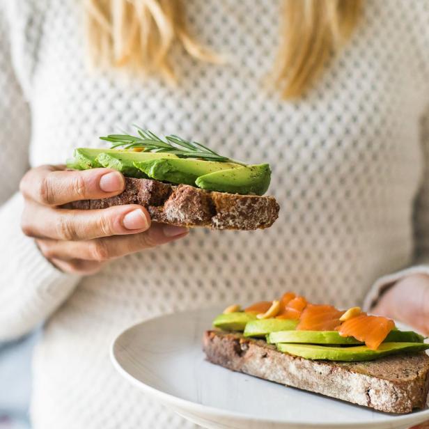 Eine Frau hält ein Avocado-Brot mit Rosmarin, während ein weiteres auf einem Teller liegt.