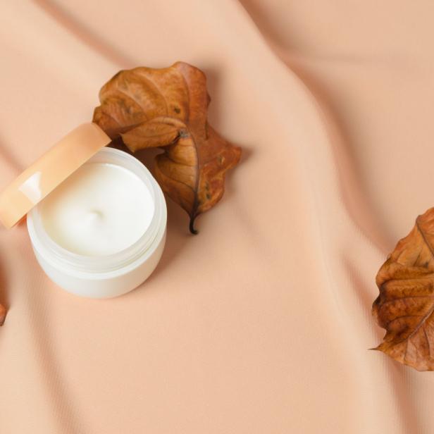 Eine geöffnete Dose mit Hautcreme liegt neben trockenen Blättern auf einem beigen Tuch.
