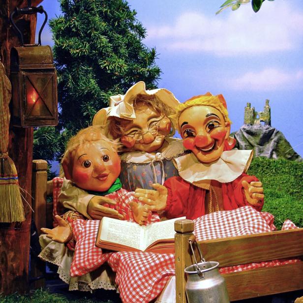 Drei Puppen sitzen in einem Bett und lesen ein Buch vor einer gemalten Landschaft.