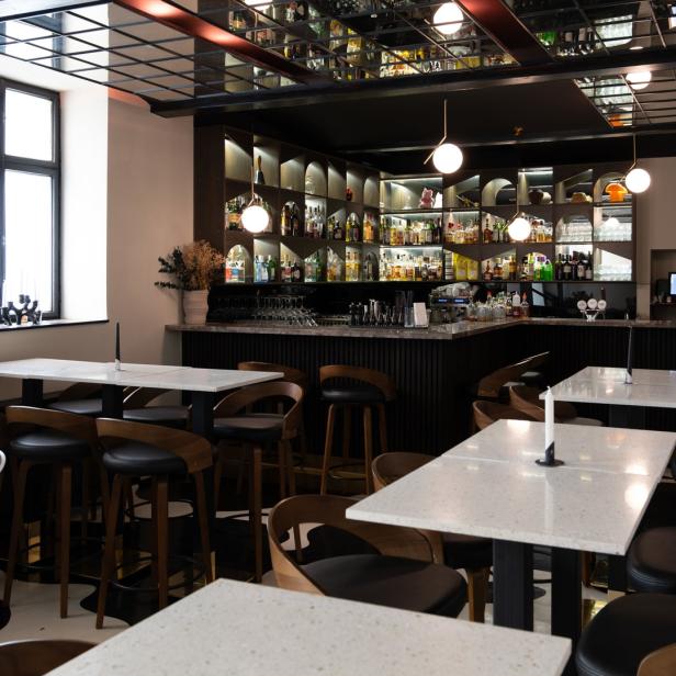 Innenansicht einer Bar mit Tischen, Stühlen und einer beleuchteten Bar im Hintergrund.