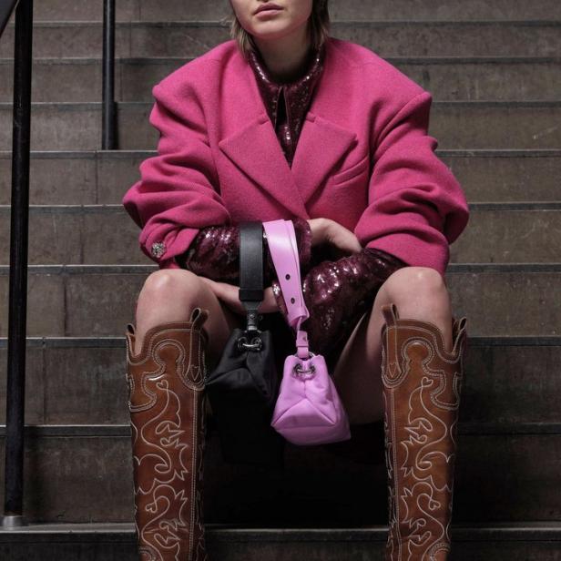 Eine Frau in Cowboystiefeln und pinkfarbenem Blazer sitzt auf einer Treppe und hält zwei Handtaschen.