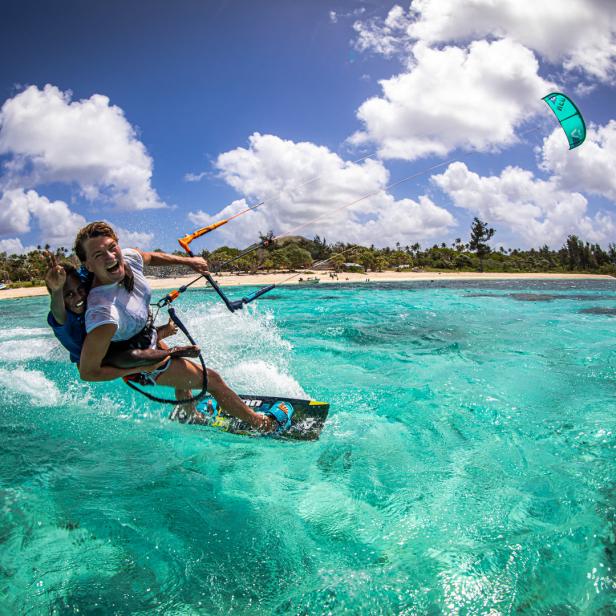 Zwei Personen beim Kitesurfen auf türkisfarbenem Wasser vor einer tropischen Küste.