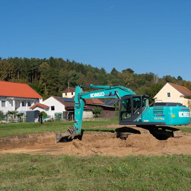 Ein türkisfarbener Bagger hebt Erde auf einer Baustelle vor Wohnhäusern und Bäumen aus.