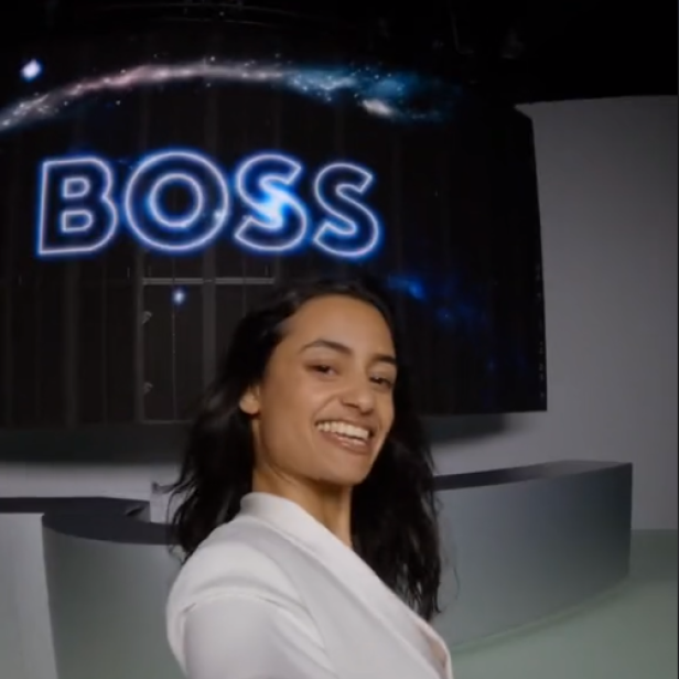 Eine Frau im weißen Blazer lächelt vor einem Bildschirm mit dem BOSS-Logo.