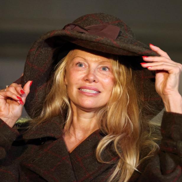 Pamela Anderson mit blonden Haaren und rotem Nagellack trägt einen braunen Hut und Mantel.
