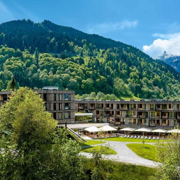 Ein modernes Hotel liegt vor einer bewaldeten Bergkulisse.