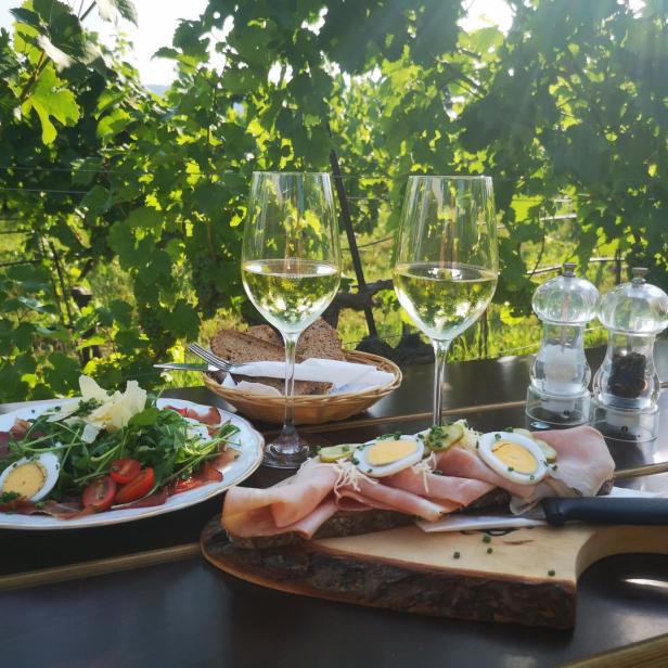 Auf einem Tisch im Weinberg stehen Wein, Brot, Wurst und Salat bereit.