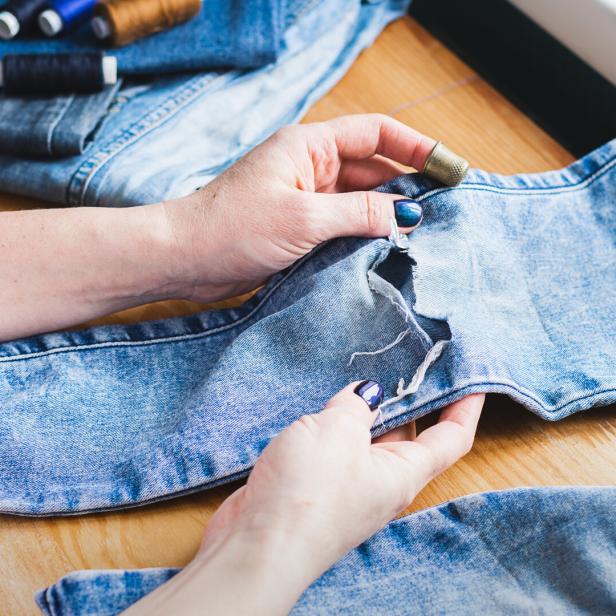 A woman is repairing ripped blue jeans. The concept of economical reuse of things. Homemade needlework.