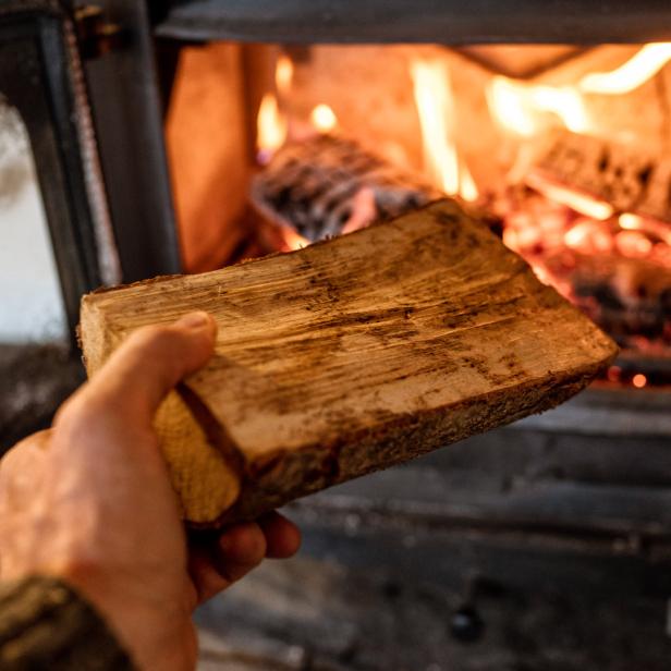 Eine Hand legt ein Holzscheit in einen brennenden Kamin.