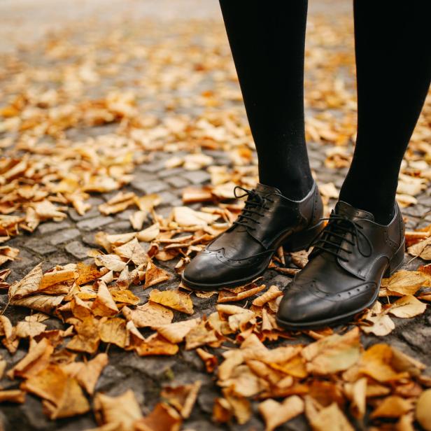 Füße in schwarzen Schuhen und Strümpfen stehen auf einem mit Herbstlaub bedeckten Weg.
