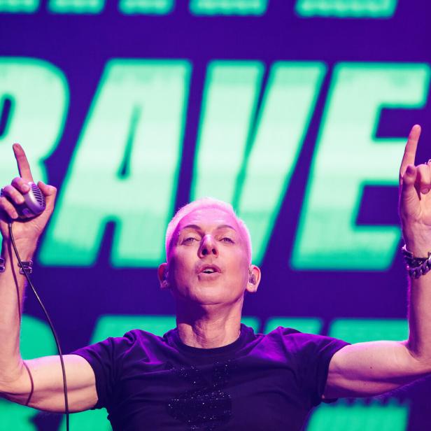 H. P. Baxxter mit erhobenen Händen steht vor einer großen „RAVE“-Leuchtschrift.