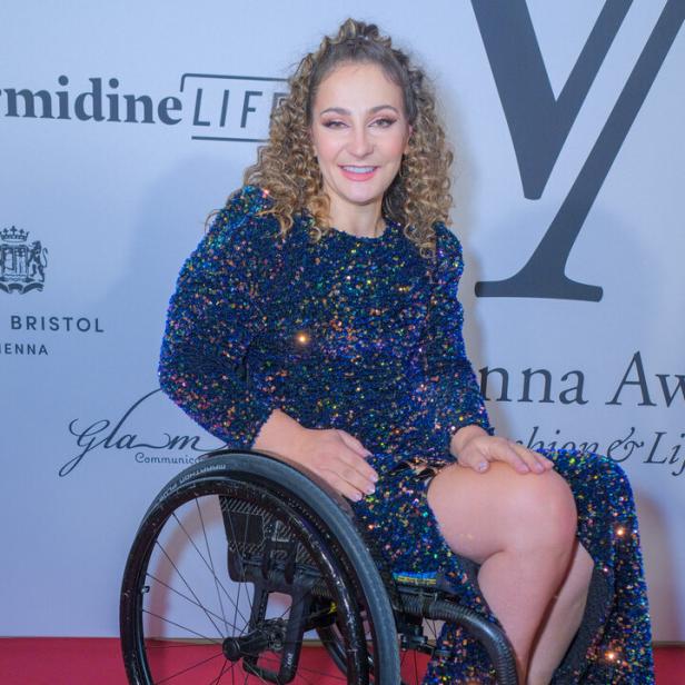 Kristina Vogel mit lockigem Haar sitzt in einem Rollstuhl und trägt ein blaues, glitzerndes Abendkleid vor einer Sponsorenwand.