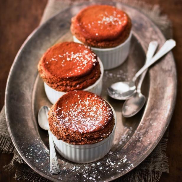 Drei Schokoladensoufflés in weißen Förmchen auf einem silbernen Tablett mit Löffeln.