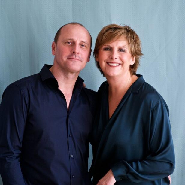 Ein Mann und eine Frau stehen lächelnd vor einem hellblauen Hintergrund.