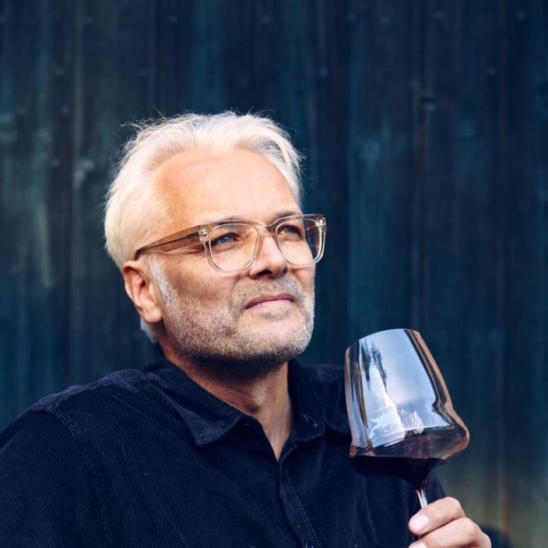 Ein Mann mit Brille betrachtet ein Glas Rotwein vor einer dunklen Holzwand.