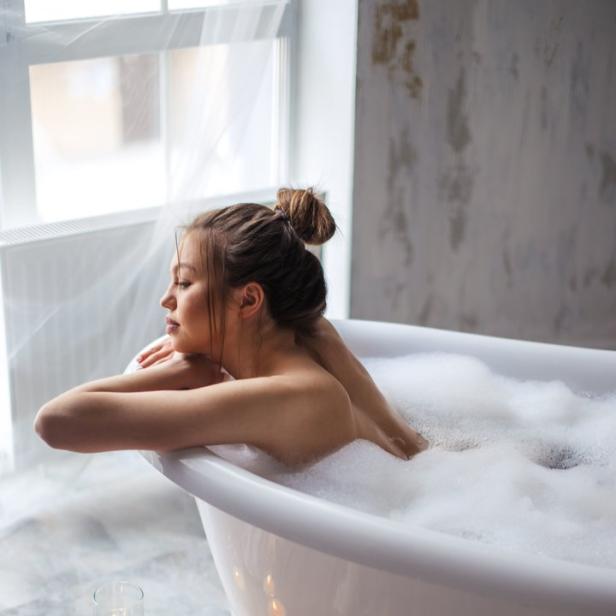 Eine Frau entspannt in einer mit Schaum gefüllten Badewanne bei Kerzenlicht.