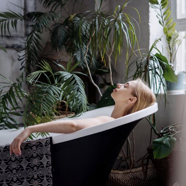 Eine blonde Frau entspannt in einer Badewanne, umgeben von Zimmerpflanzen.