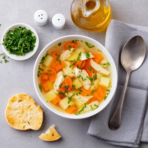 Eine Schale Hühnersuppe mit Gemüse und frischen Kräutern, daneben Brot, Kräuter und ein Löffel auf Serviette.