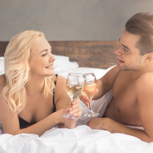 Ein Paar liegt im Bett und stößt mit Weißwein an.