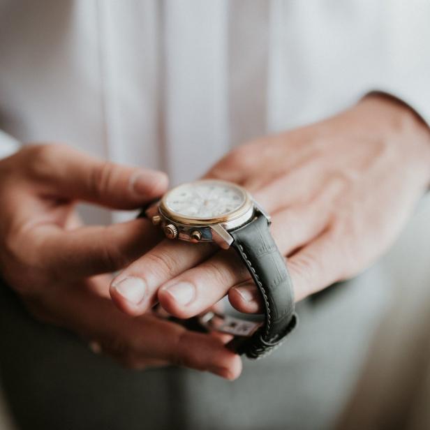 Ein Mann hält eine goldene Armbanduhr mit schwarzem Lederarmband in seinen Händen.