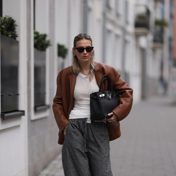 Eine Frau mit Sonnenbrille, brauner Jacke und grauer Hose geht auf dem Bürgersteig.