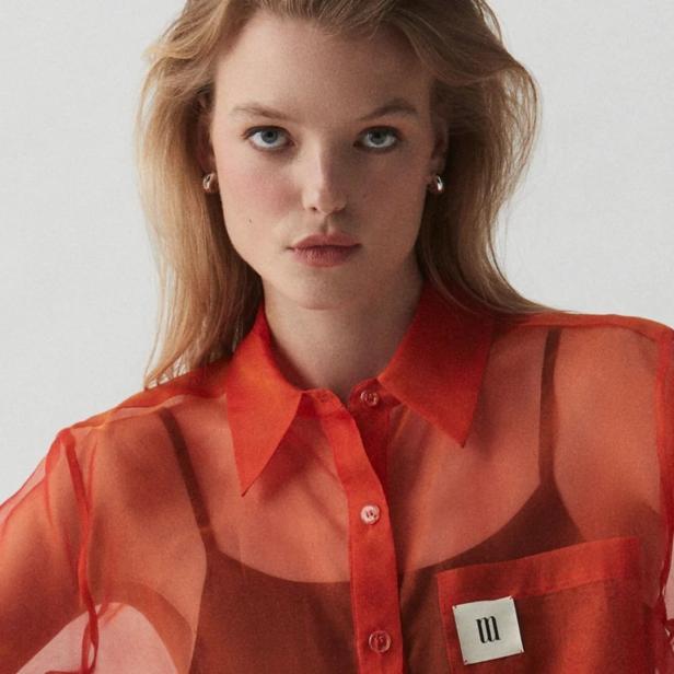 Eine Frau mit blonden Haaren trägt eine orange, transparente Bluse.