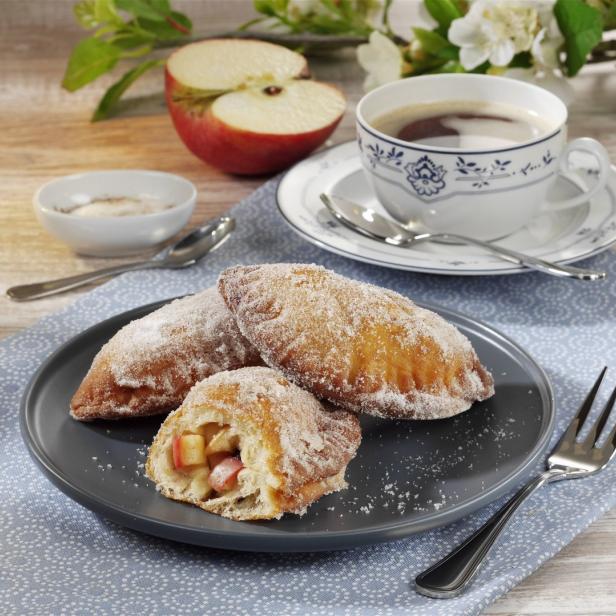 Apfeltaschen mit Puderzucker auf einem Teller, dazu eine Tasse Kaffee und eine halbe Apfel.