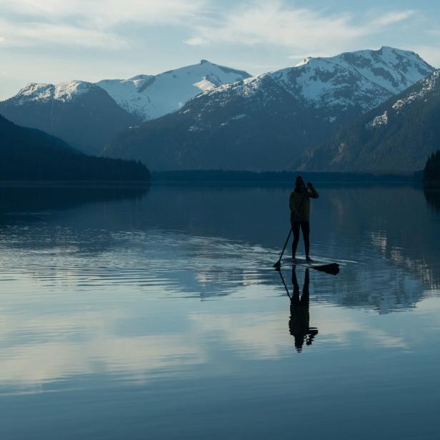 Eine Person steht auf einem Paddleboard auf einem ruhigen See vor einer Bergkulisse.