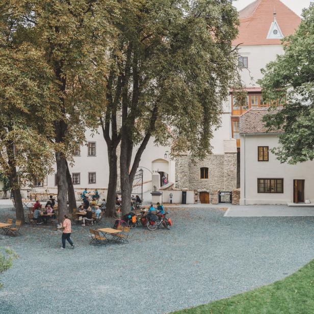 Ein Innenhof mit Tischen und Stühlen, umgeben von alten Gebäuden und Bäumen.