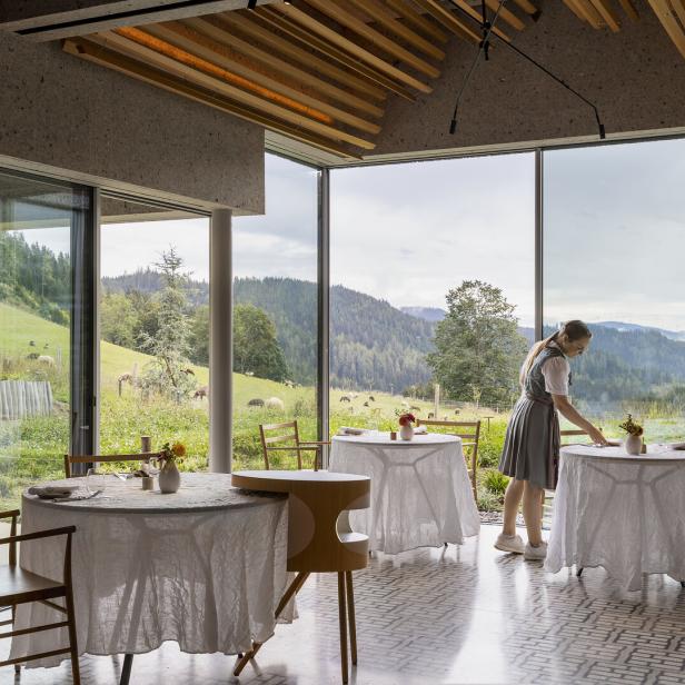 Eine Frau im Dirndl deckt Tische in einem Restaurant mit Blick auf eine grüne Landschaft.
