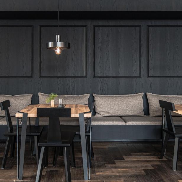 Ein Restaurantbereich mit Holztischen, schwarzen Stühlen und einer gepolsterten Sitzbank vor einer dunklen Holzvertäfelung.