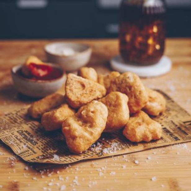 Eine Portion goldbraune Nuggets mit Dipsaucen und einem Getränk mit Strohhalmen im Hintergrund.