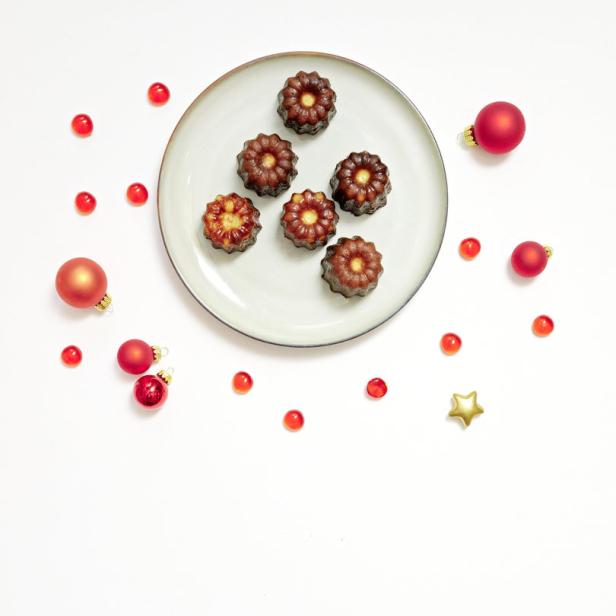 Canelés auf einem Teller, umgeben von roten Weihnachtskugeln und Dekorationen.