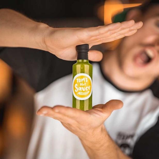 Ein Mann präsentiert aufgeregt eine Flasche „Tom's Hot Sauce“.