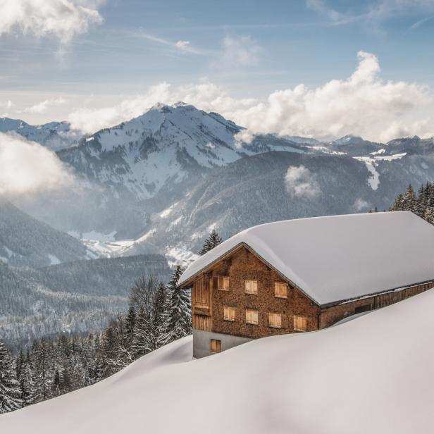 Ein schneebedecktes Holzhaus vor einer verschneiten Berglandschaft.