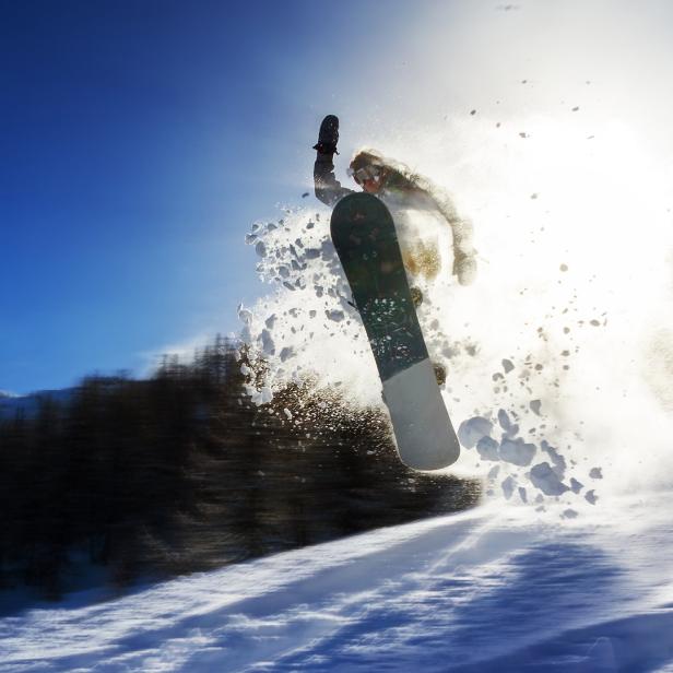 Ein Snowboarder springt mit viel Schnee in der Luft über eine Piste.