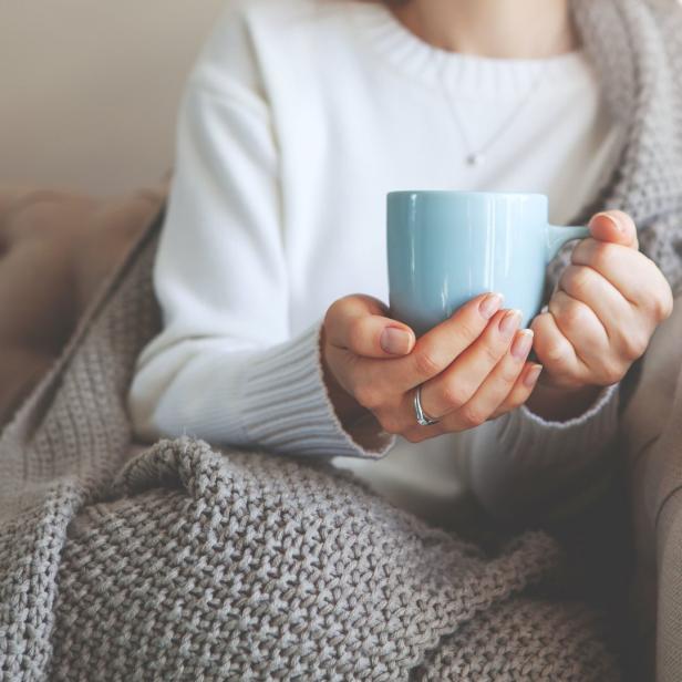 Eine Frau sitzt mit einer hellblauen Tasse und einer grauen Decke auf einem Sofa.