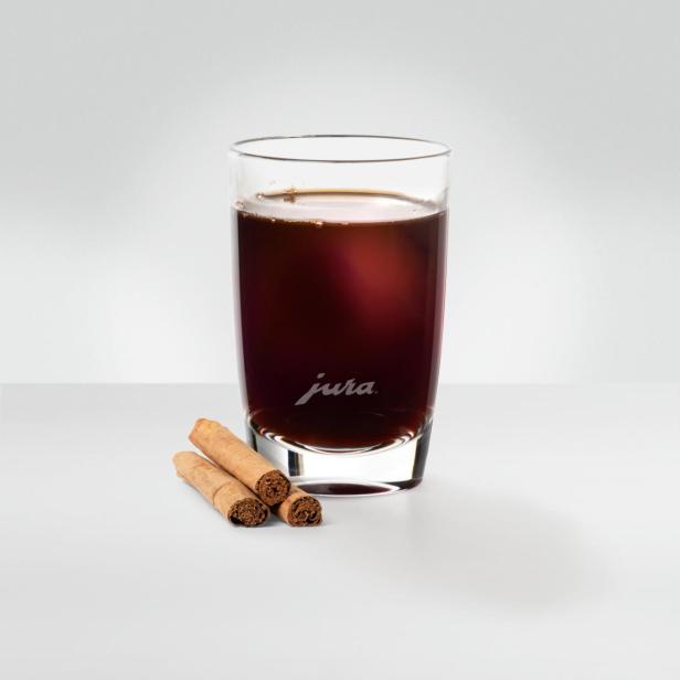 Ein Glas mit dunklem Kaffee und drei Zimtstangen auf einem weißen Tisch.