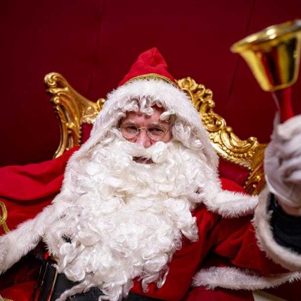 Weihnachtsmann mit Bart und Glocke in der Hand sitzt auf einem goldenen Thron.