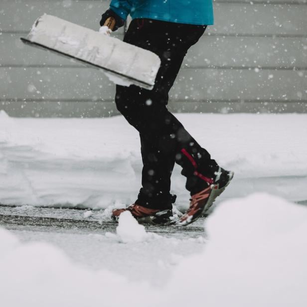 Eine Person schiebt im Schneefall Schnee mit einer Schneeschaufel beiseite.